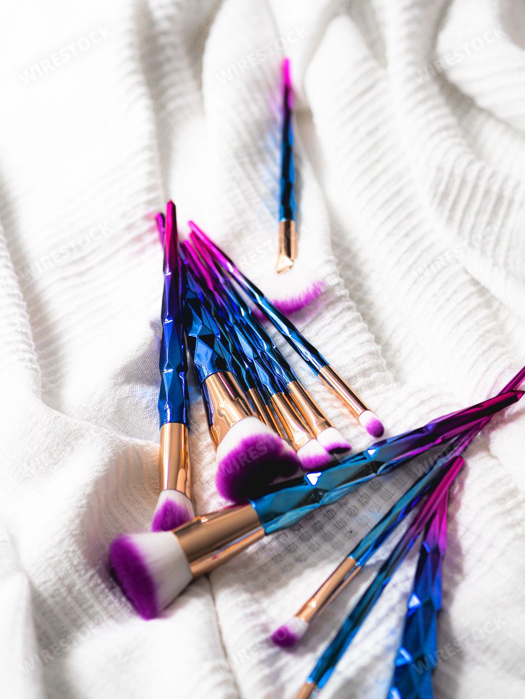 A vertical closeup shot of numerous purple and blue makeup brushes on a white cloth