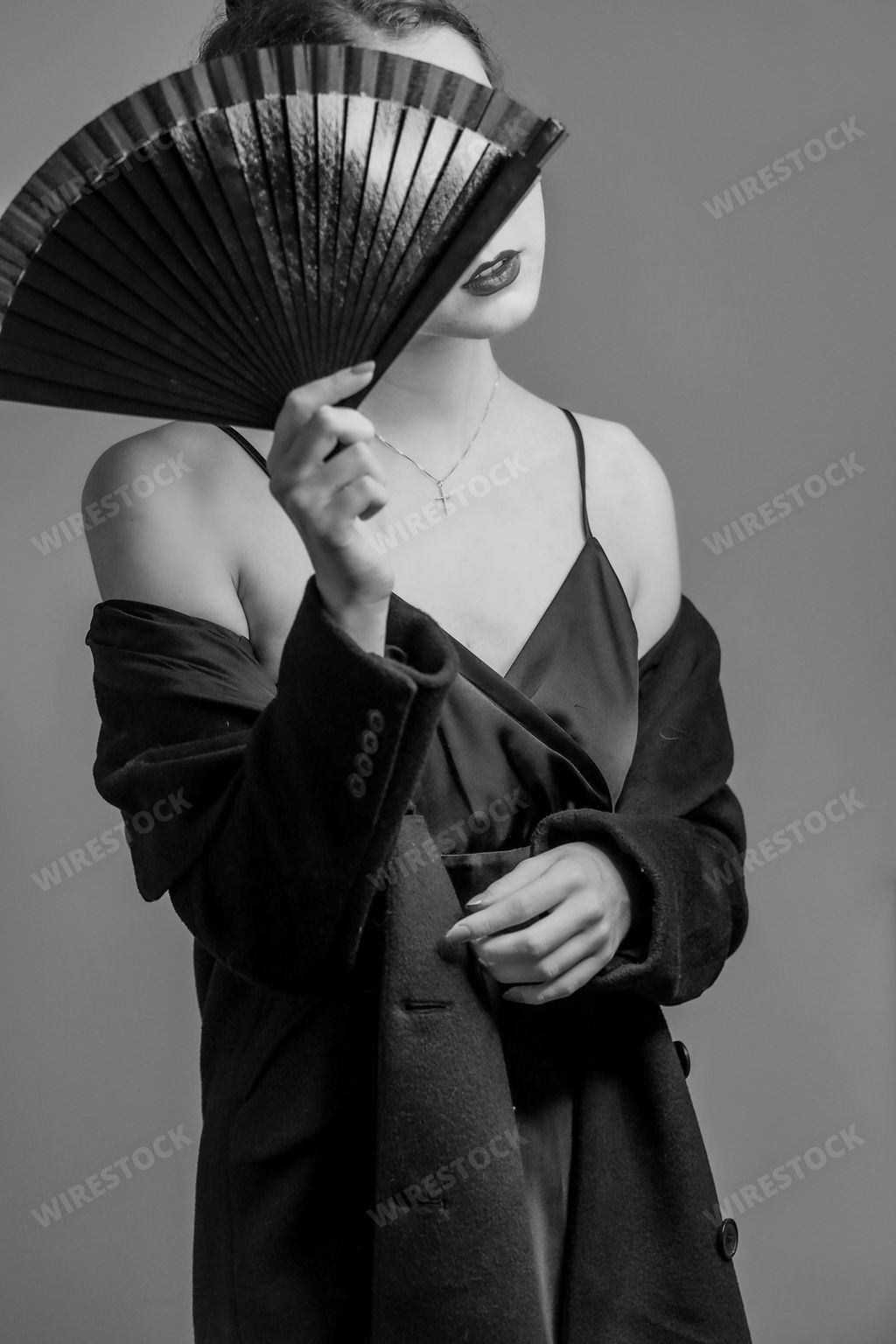 A vertical grayscale shot of an elegant female posing in a slip dress, trench coat, and a fan