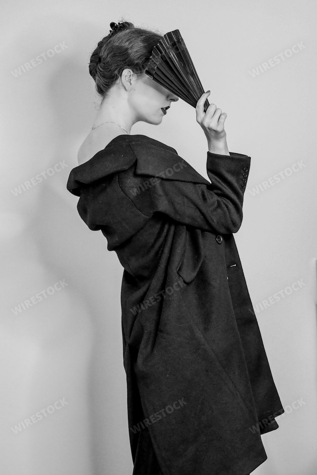 A vertical grayscale shot of an elegant female posing in a slip dress, trench coat, and a fan