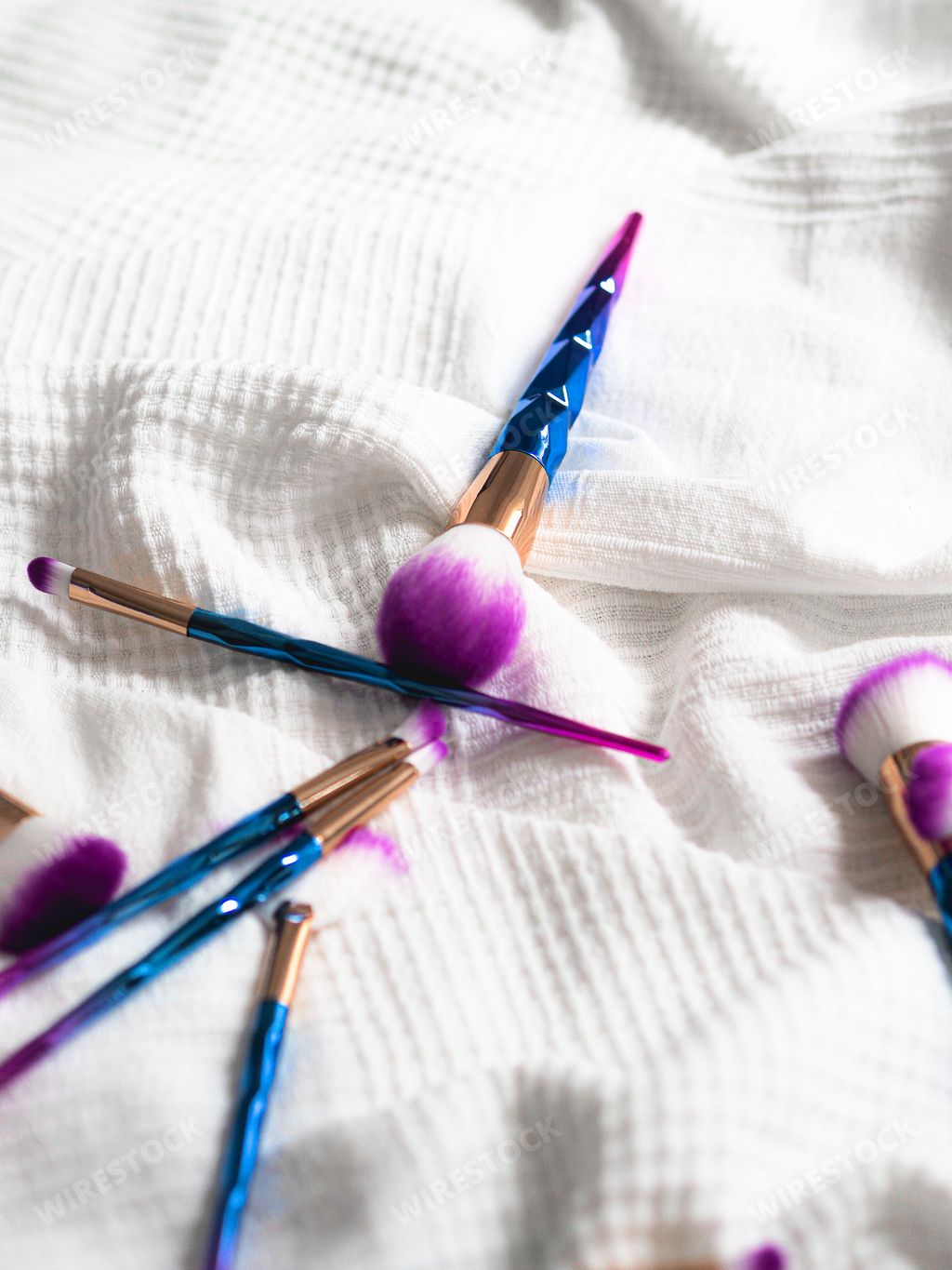 A vertical closeup shot of numerous purple and blue makeup brushes on a white cloth