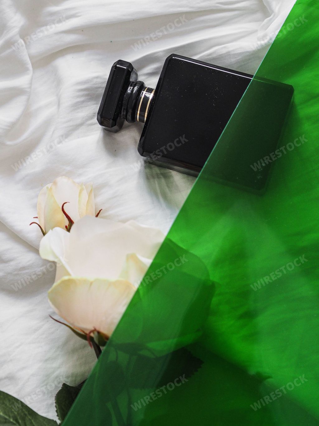 A vertical shot of a black perfume bottle on bed sheets with a white rose