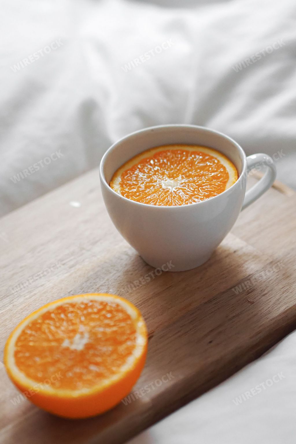 A vertical shot of a tea cup with a sliced orange in it next to a half orange on wooden board on white fabric