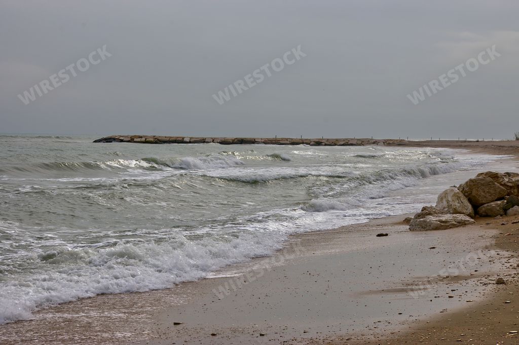 A horizontal view of foamy sea waves crashing on a sandy beach under a cloudy sky in Italy
