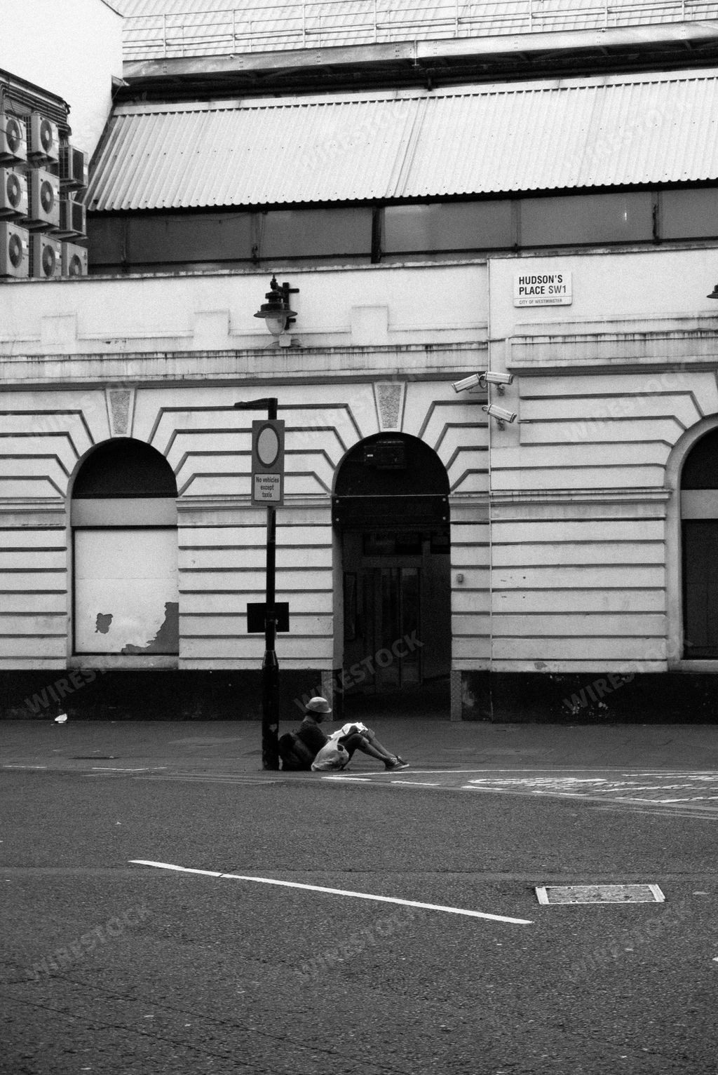 A grayscale shot of a person sitting and leaning against a post on the side of a street