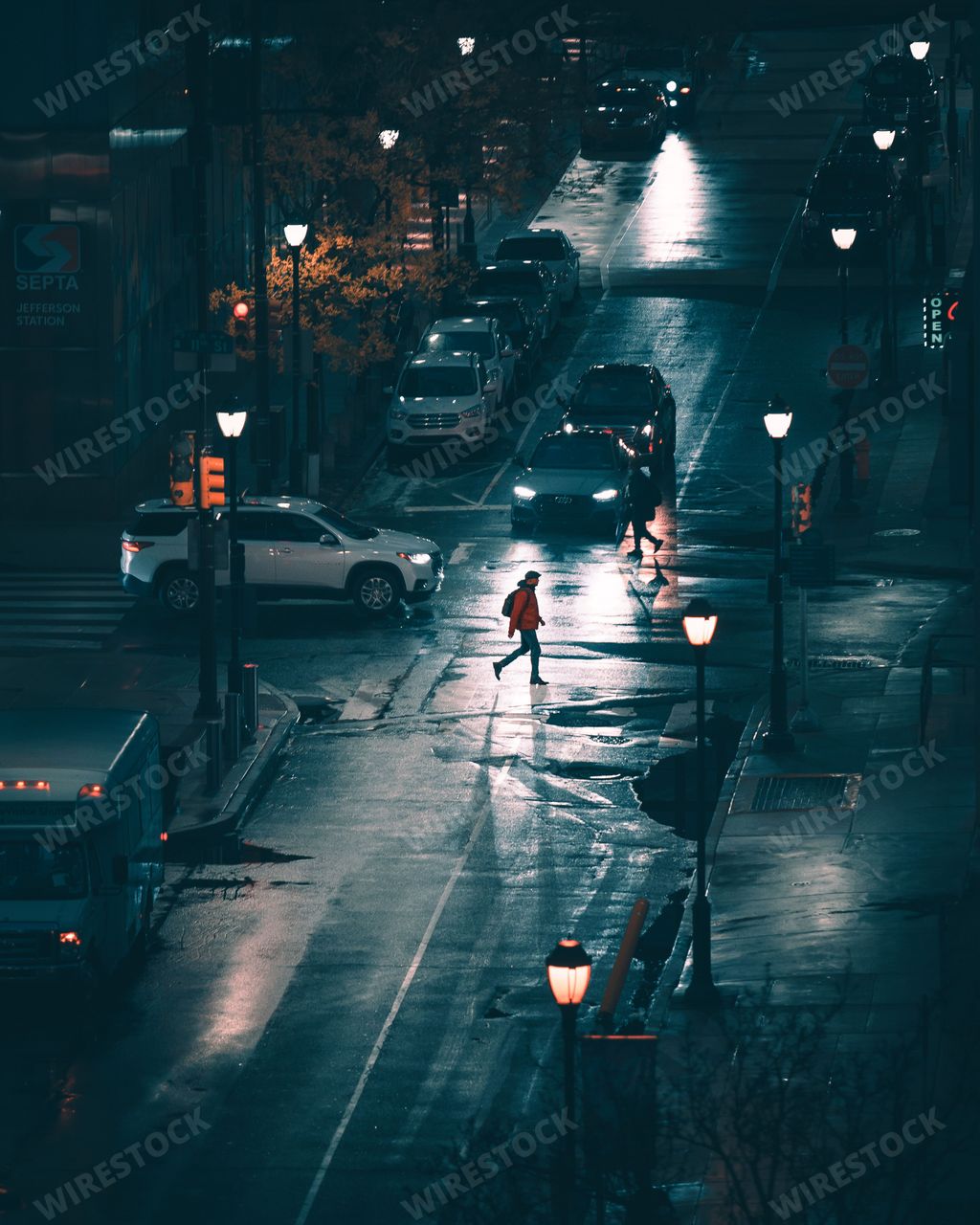 A lone figure strides down a city street illuminated by street lamps, the scene shrouded in a light rain