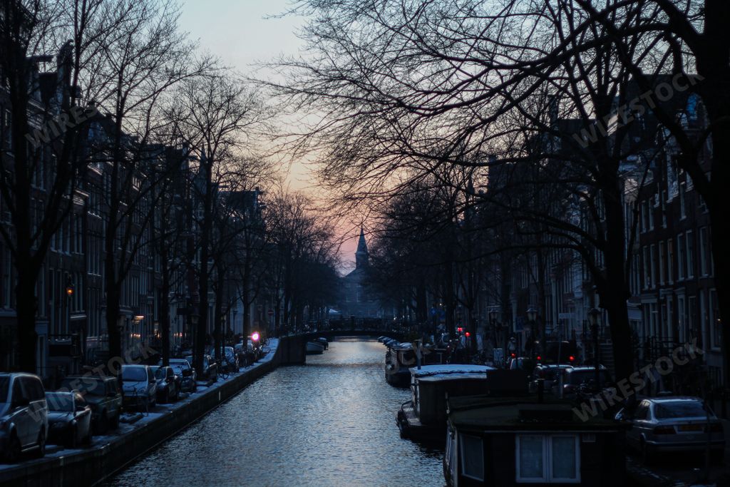 A scenic view of boats in a river flowing through Amsterdam, the Netherlands with both sides surrounded by cars and houses