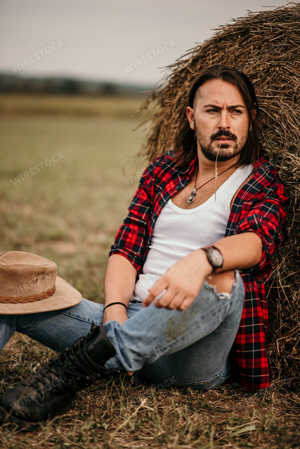 A young handsome Serbian male in a flannel shirt and ripped jeans sitting leaned on a haystack