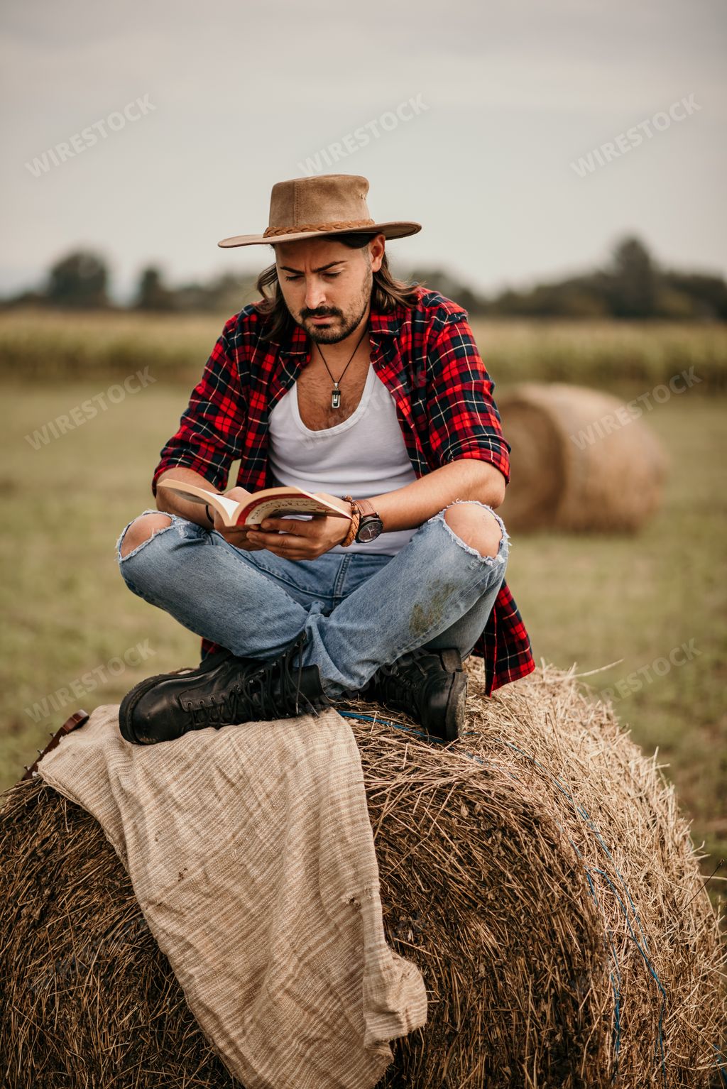 A young handsome Serbian male in a flannel shirt sitting on a haystack and reading a book