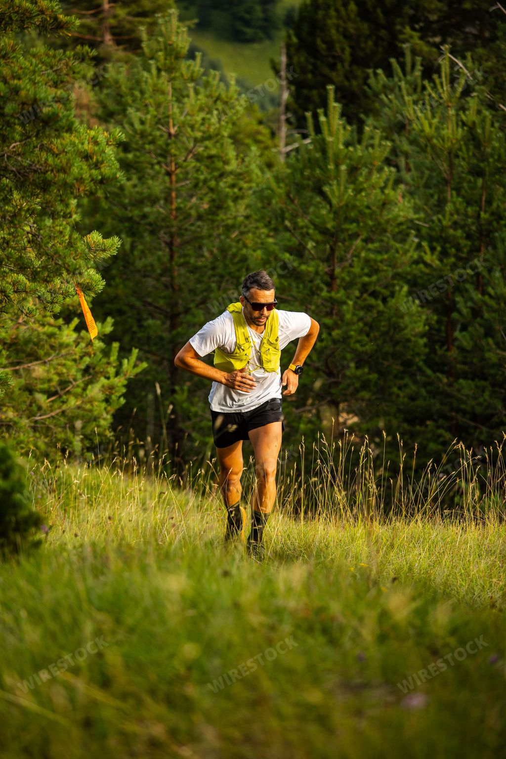 A vertical shot of a Caucasian man running through a forest trail wearing a yellow vest and sunglasses on a sunny day