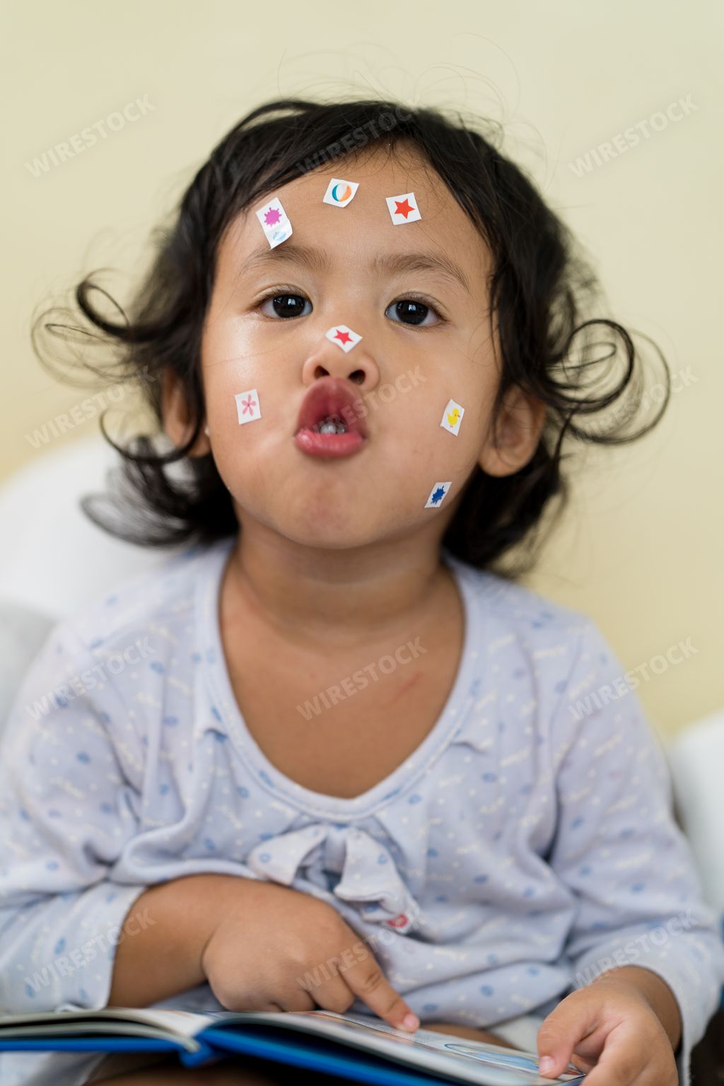 A vertical shot of a cute Southeast Asian toddler girl with stickers on her face and an open book