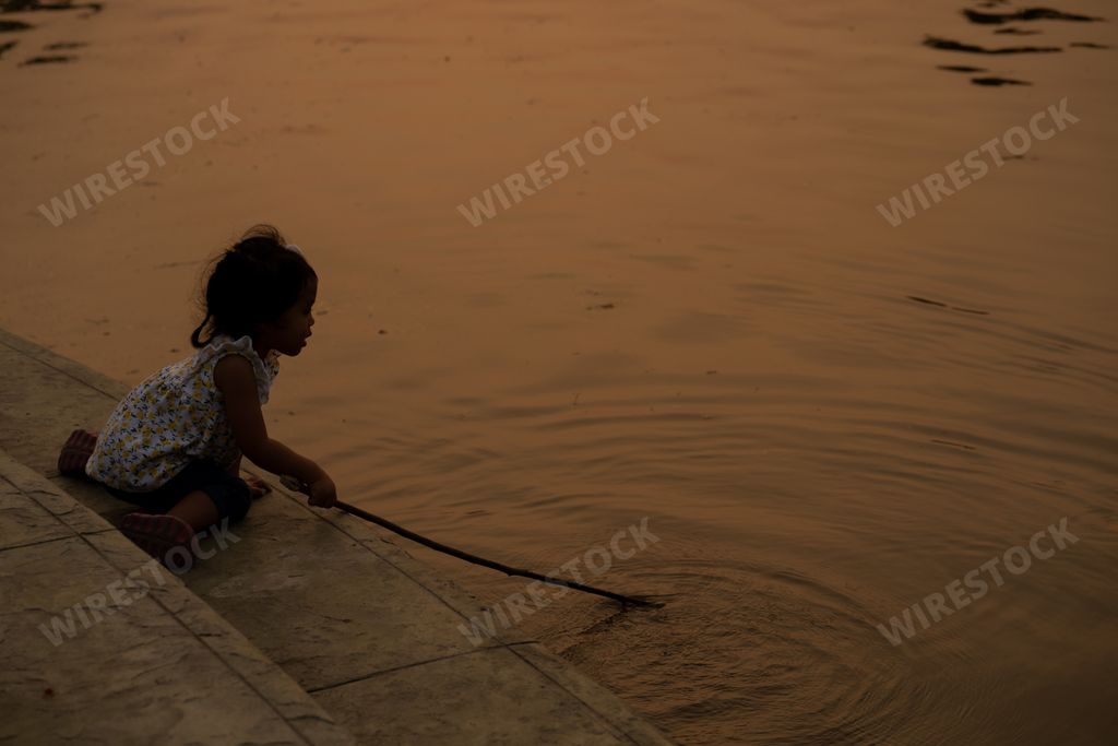 A little Thai girl siting holding wooden stick playing near the pond at sunset