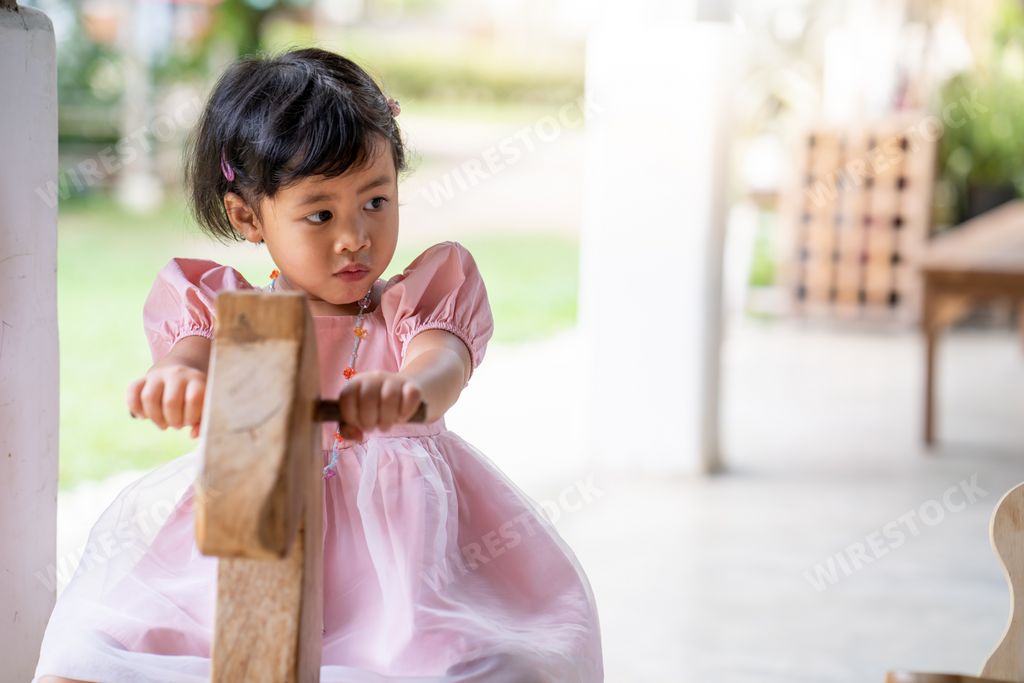 A shallow focus of a young Southeast Asian girl in a pink dress riding a wooden rocking horse at a patio