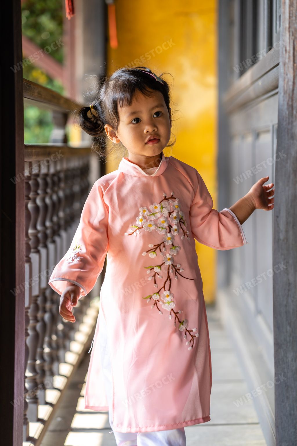 A vertical shot of a cute Vietnamese little girl in a pink ao dai walking on an old wooden balcony