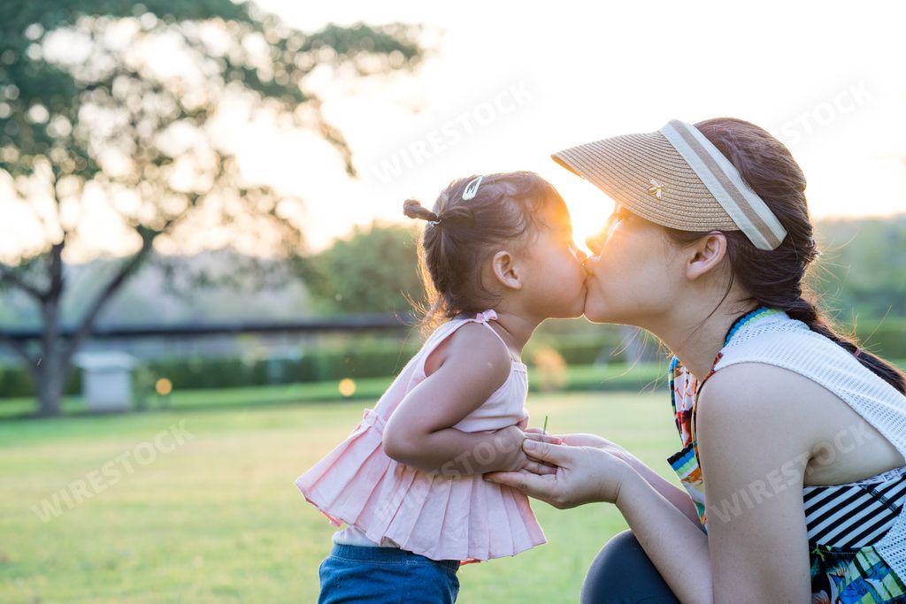 a thai mother kissing child with holding hands in green field under sunlight