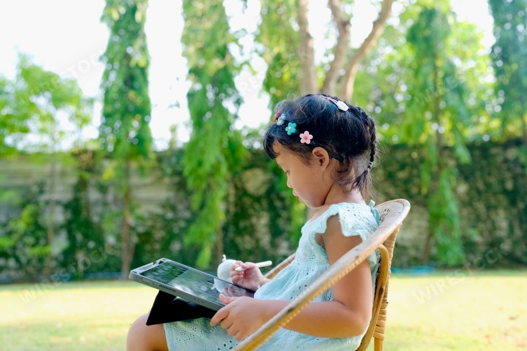 An adorable little Thai girl sitting in a lush green park and drawing on her tablet on a sunny day