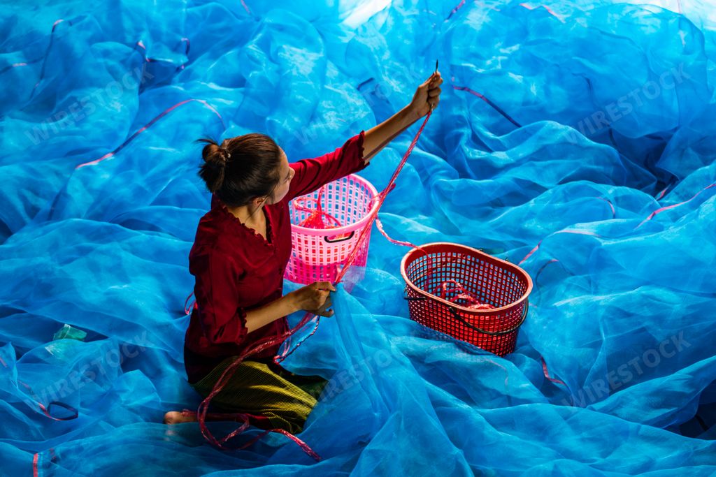 An overhead shot of a Southeast Asian woman sewing a blue fishnet as a livelihood