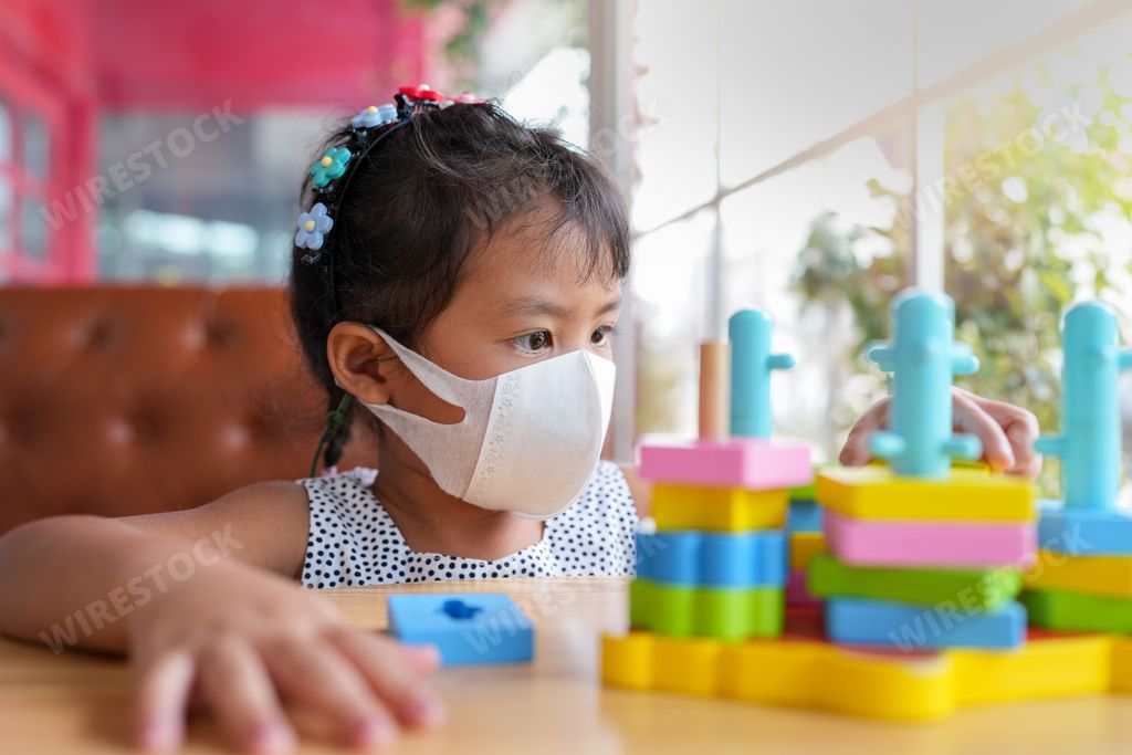 A southeast-asian girl wearing a protective face mask sitting at a table, playing with colorful building blocks