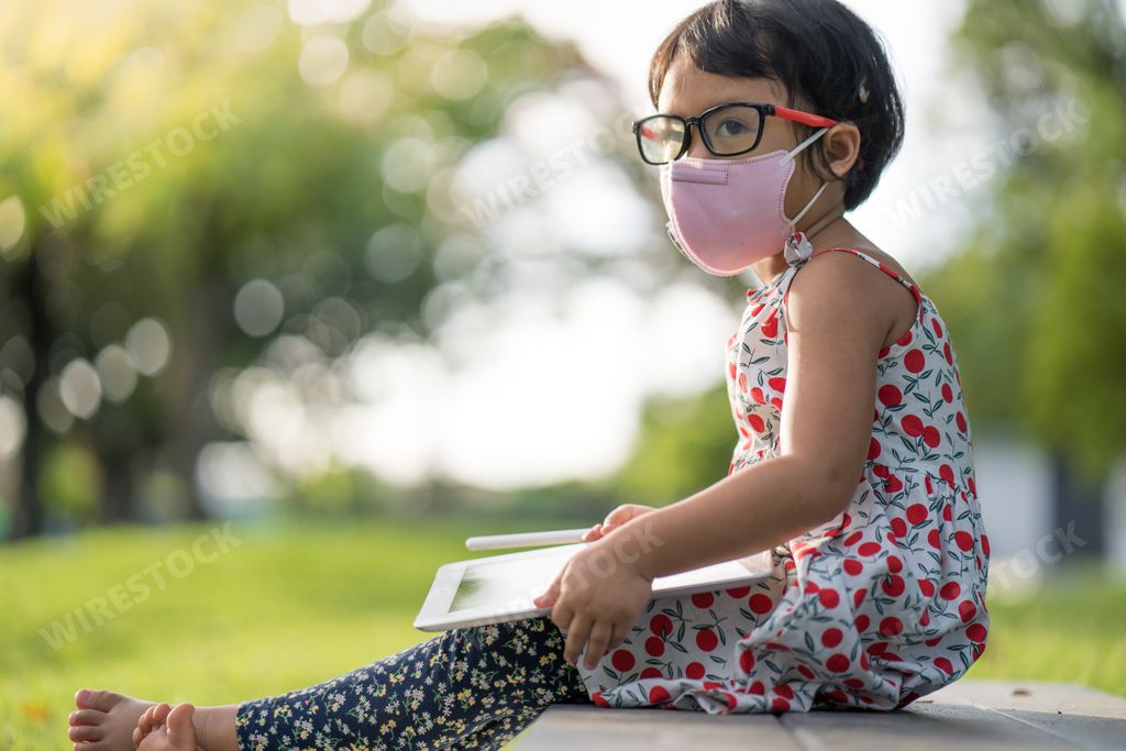 A cute Asian small girl in a facial mask sitting on the grass and drawing on a tablet