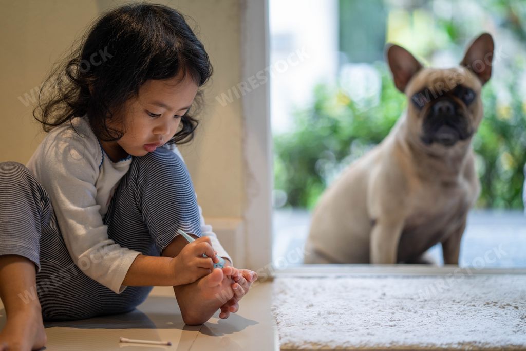 A cute little girl painting her toenails with her dog indoors.
