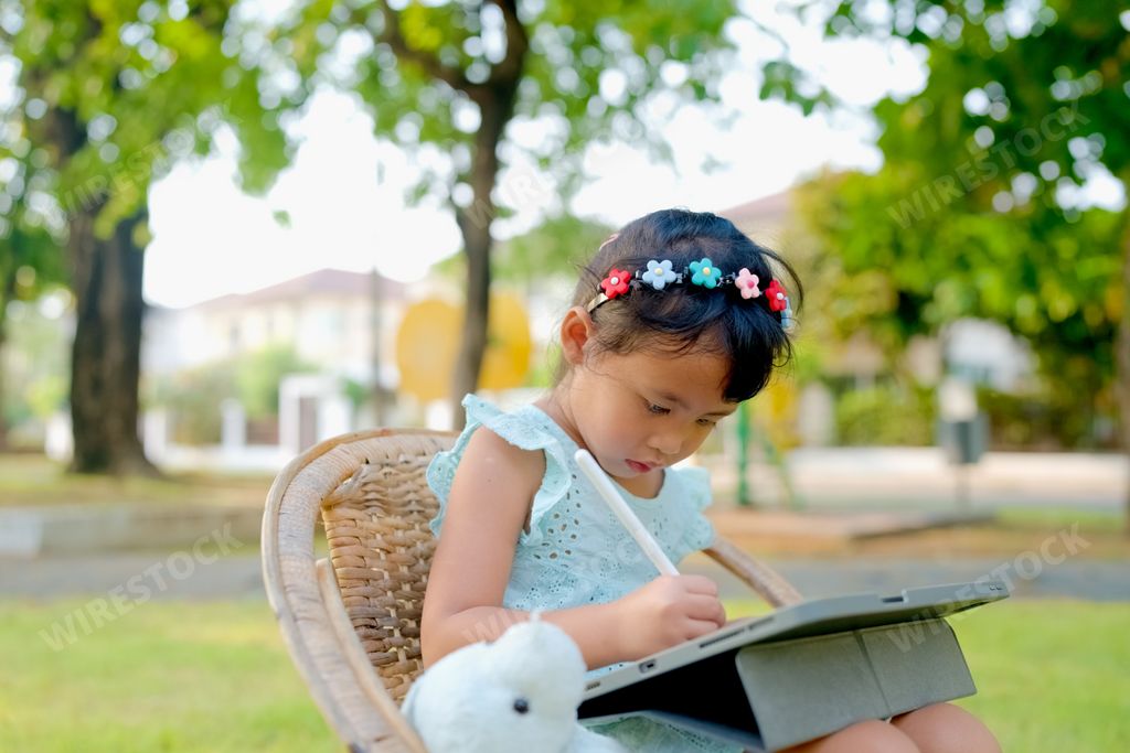 An adorable little Thai girl sitting in a bright green park and drawing on her tablet with her stuffed bunny beside her