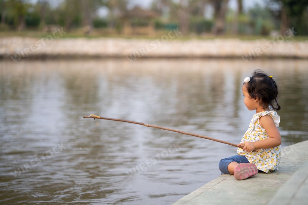 A closeup of a little girl sitting near the water with a long stick in her hand.