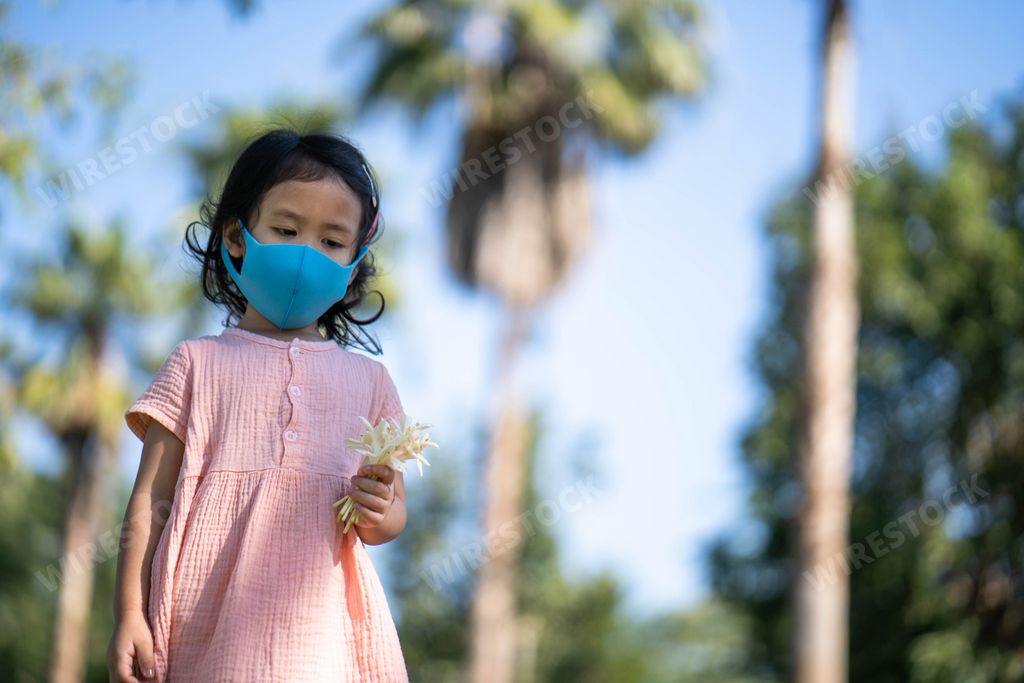 A Thai cute little girl wearing a face mask holding white flowers while  walking outdoor