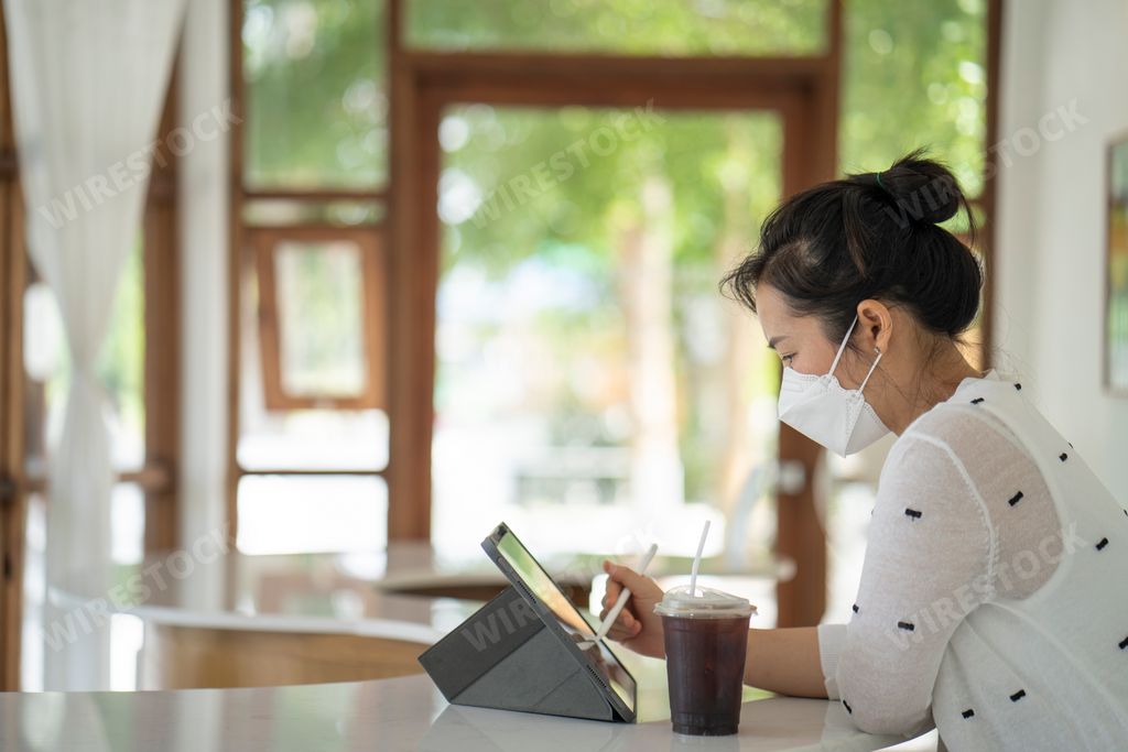 An Asian woman working on her tablet in a cafe with a face mask on