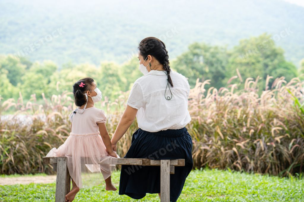 A young mother and her child from Thailand in masks on the bench in the garden during the coronavirus