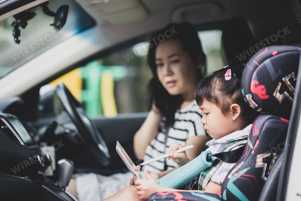 The Southeast Asian female kid in school uniform using a mobile tablet beside her mother in the car
