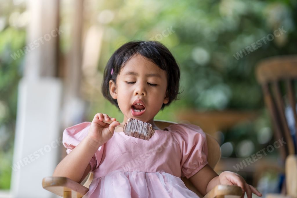 A shallow focus of a young Southeast Asian girl eating a popsicle while sitting on a wooden chair