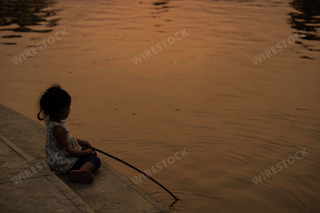 A little Thai girl sitting holding a wooden stick playing near the pond at sunset