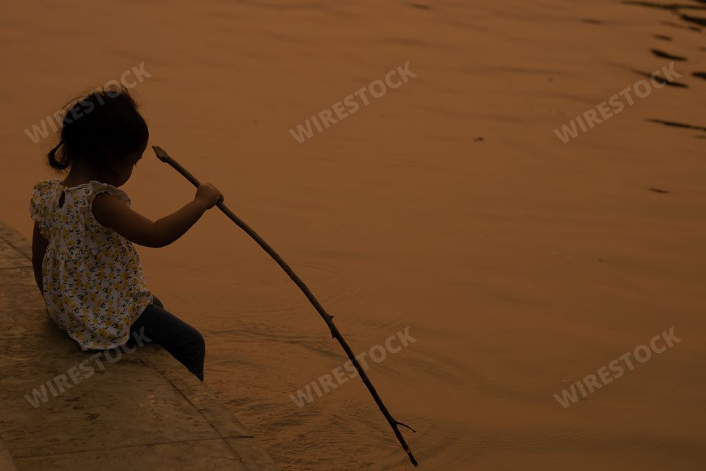 A Thai cute little girl holding a wooden stick playing near a pond during sunset