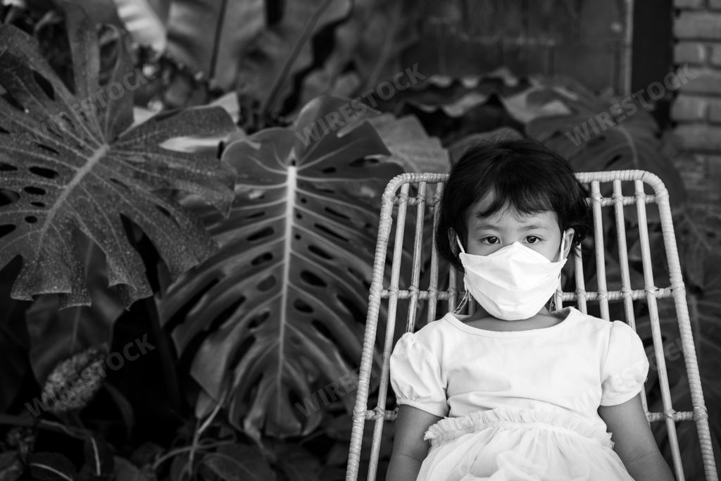 A grayscale shot of a little Asian girl wearing a face mask sitting on a chair
