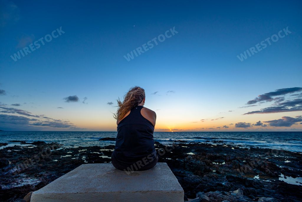 A back view of a female sitting on a shore and looking at calm sea during sunset