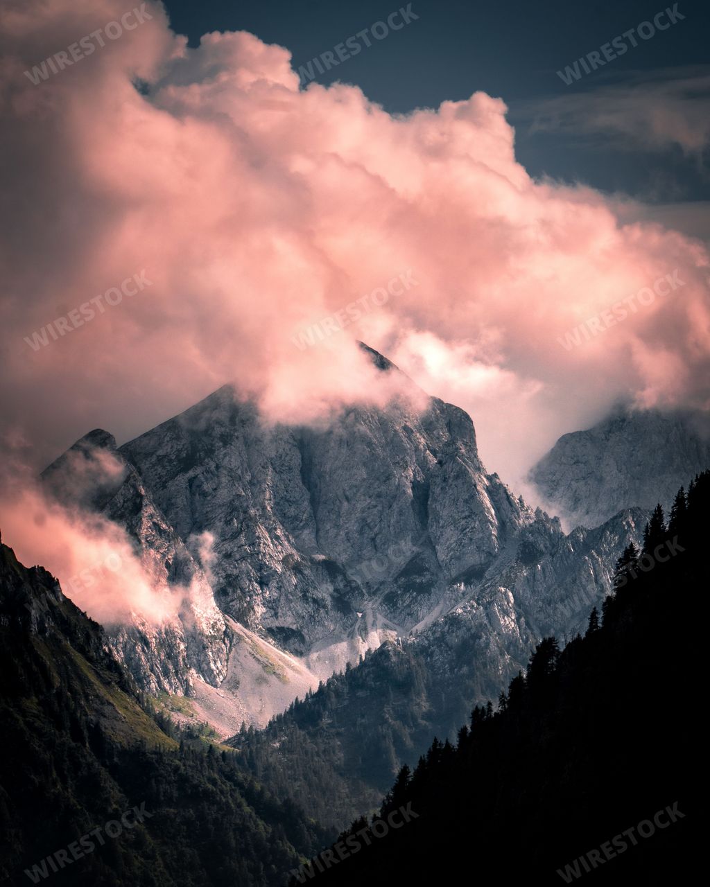 A vertical aerial view of a rocky peak of a green mountain range covered with clouds at sunset