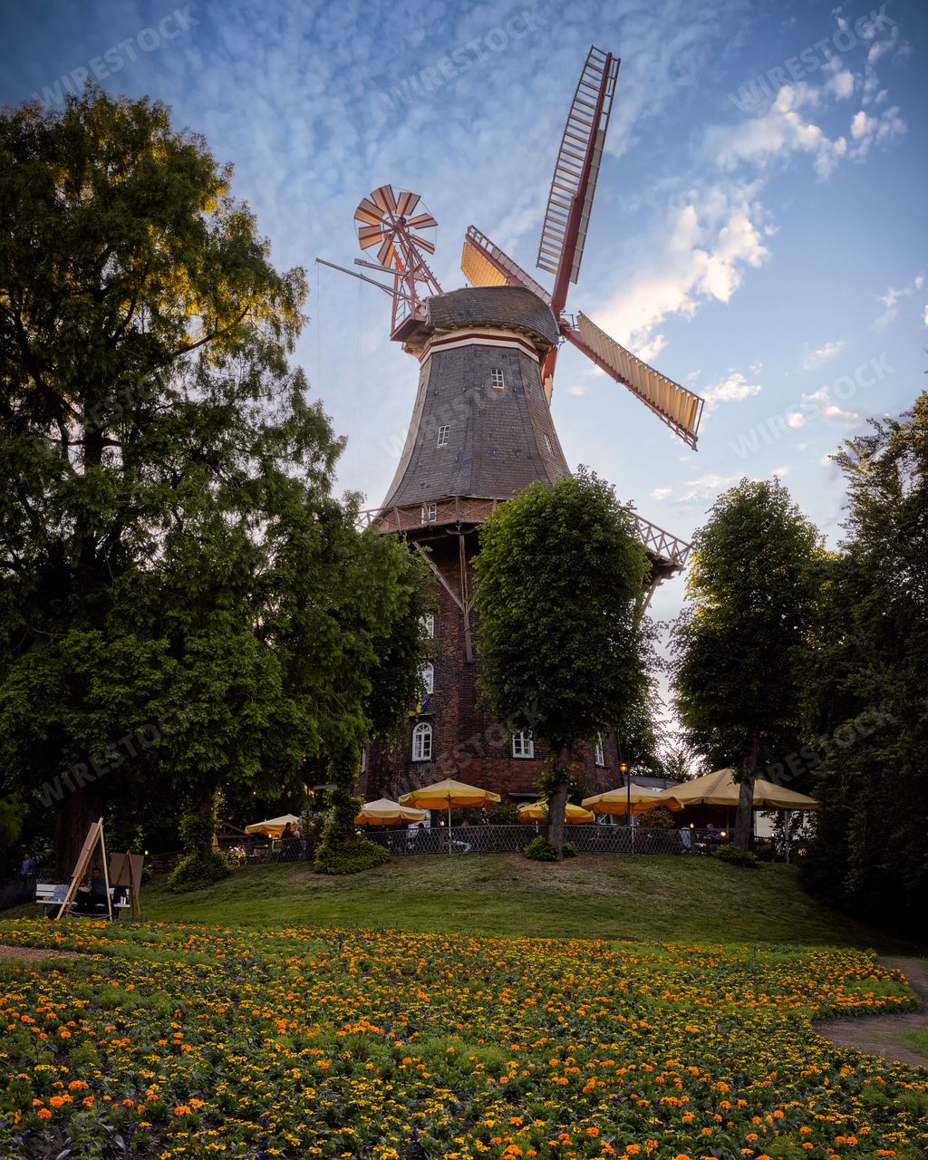 A picturesque windmill surrounded by lush greenery and vibrant flowers under a partly cloudy sky in Bremen, Germany.