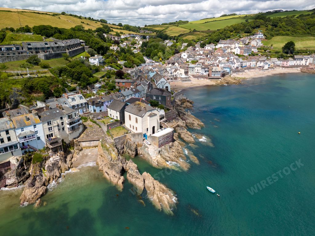 Aerial view of a coastal village with houses on cliffs and a beach, surrounded by green hills and a blue sea.