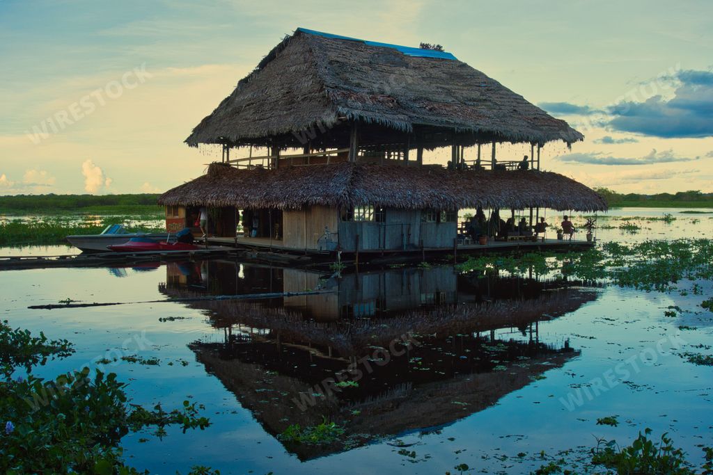 A picturesque thatched roof cottage situated on a tranquil body of water, with various vehicles parked in front of it