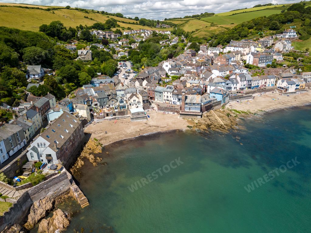 Aerial view of a coastal village with colorful houses, green hills, and a sandy beach in Cornwall, UK