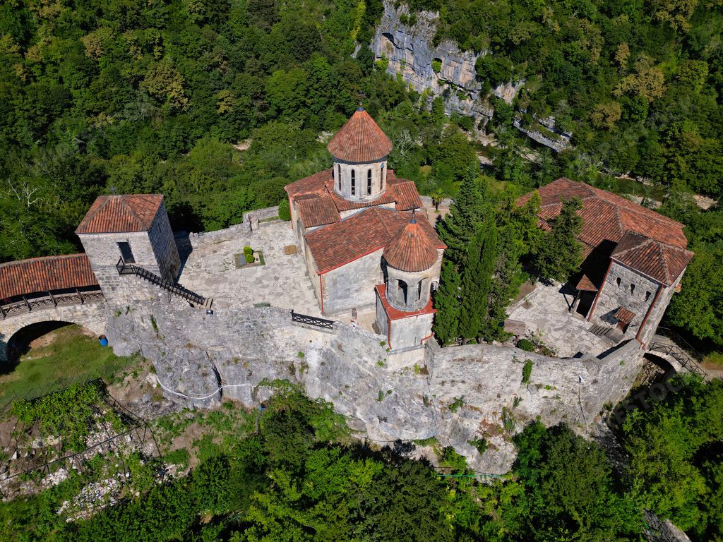 A picturesque castle with a red-tiled roof and two chimneys atop stone walls, situated on a hilltop overlooking the surrounding countryside