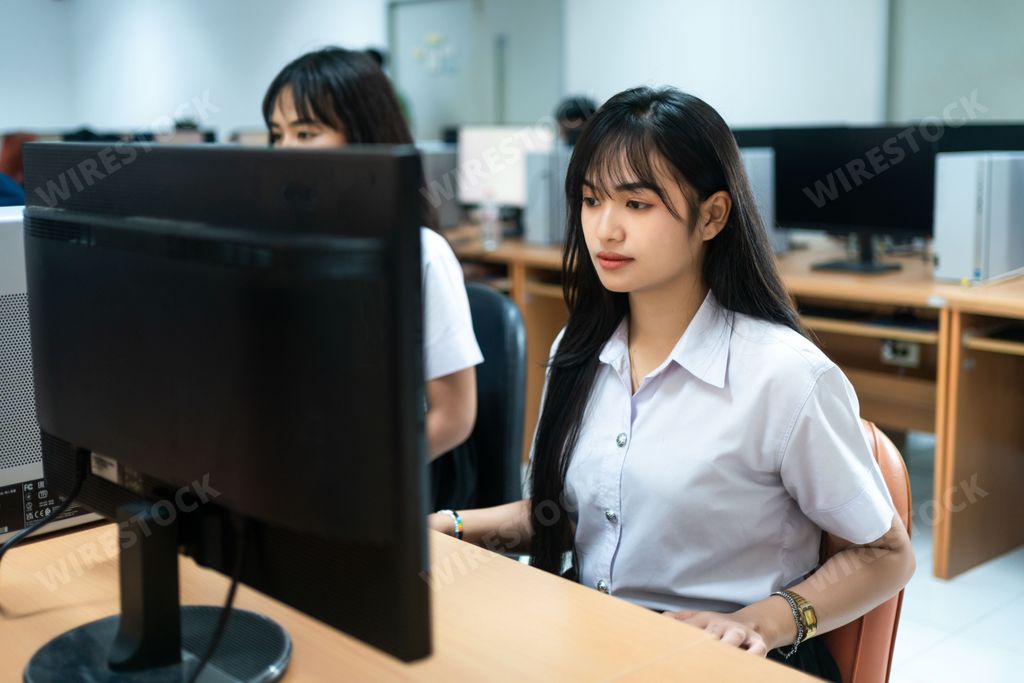 Two young Asian female students sitting at a desk in front of a laptop computer, smiling and looking at the screen