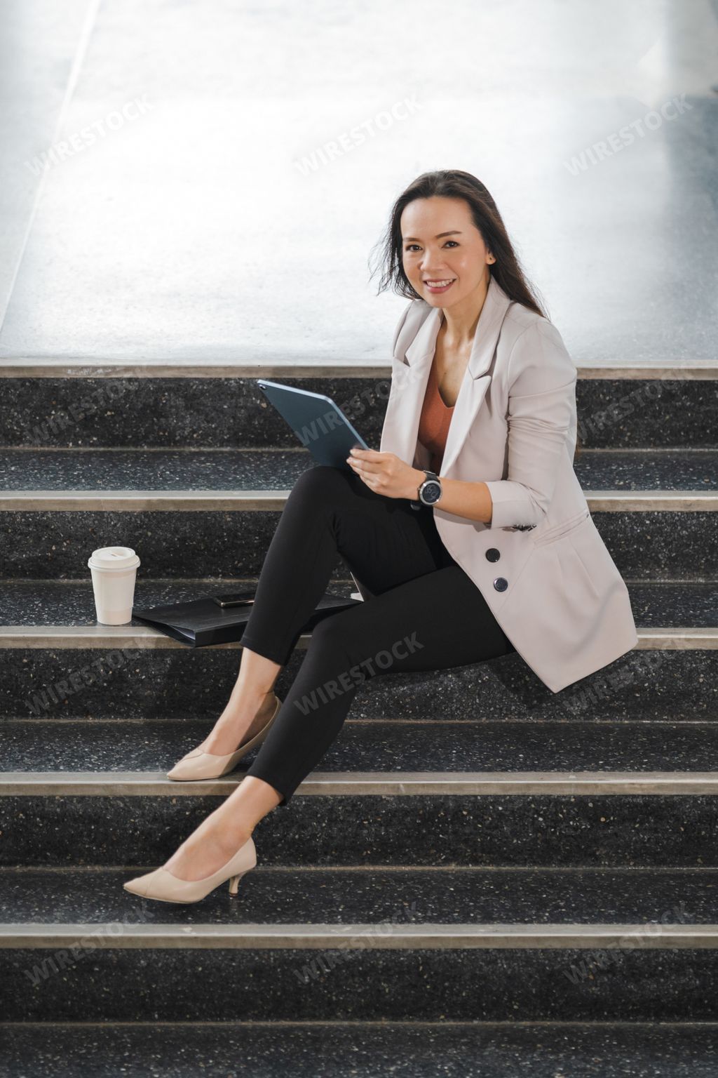 A cheerful Thai young businesswoman sitting on the stairs working on her tablet in the office
