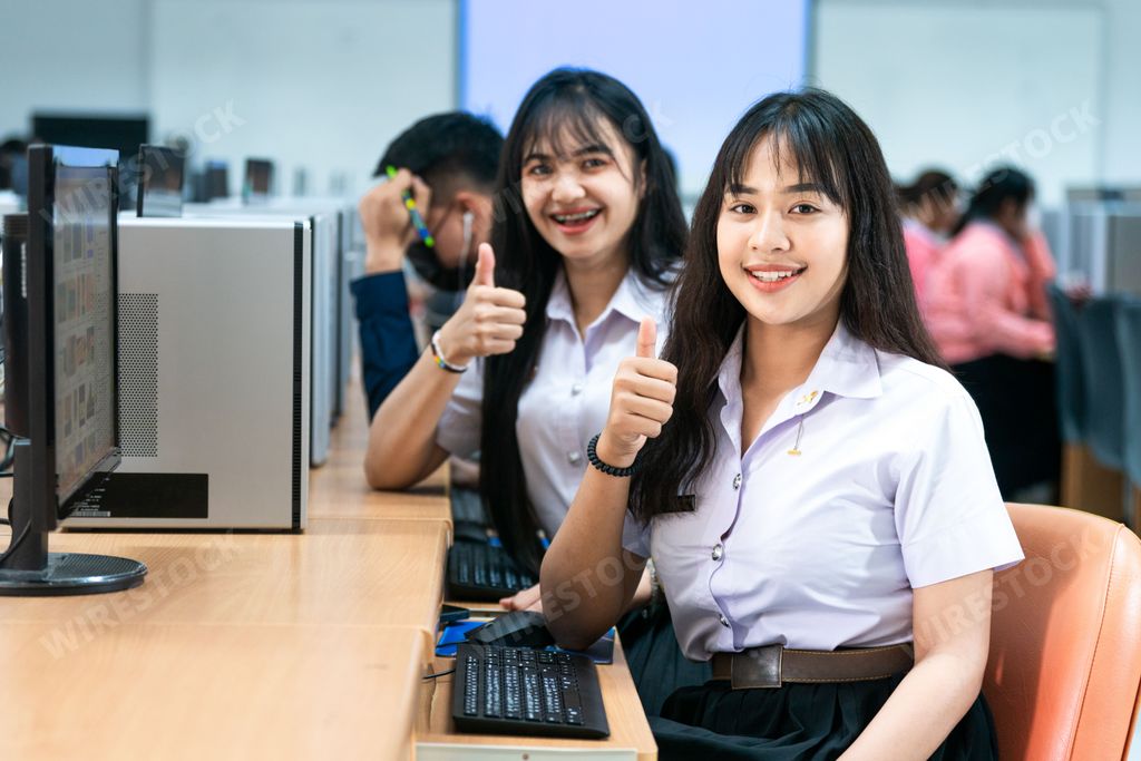 Two young Asian female students seated at a desk in front of a laptop computer, with thumbs up