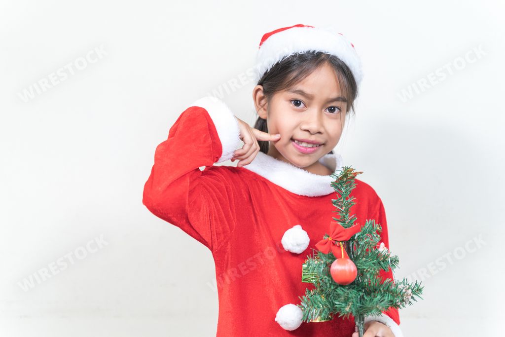 Little cute Thai girl with Santa costume touching her cheek and holding a Christmas tree on a white background.