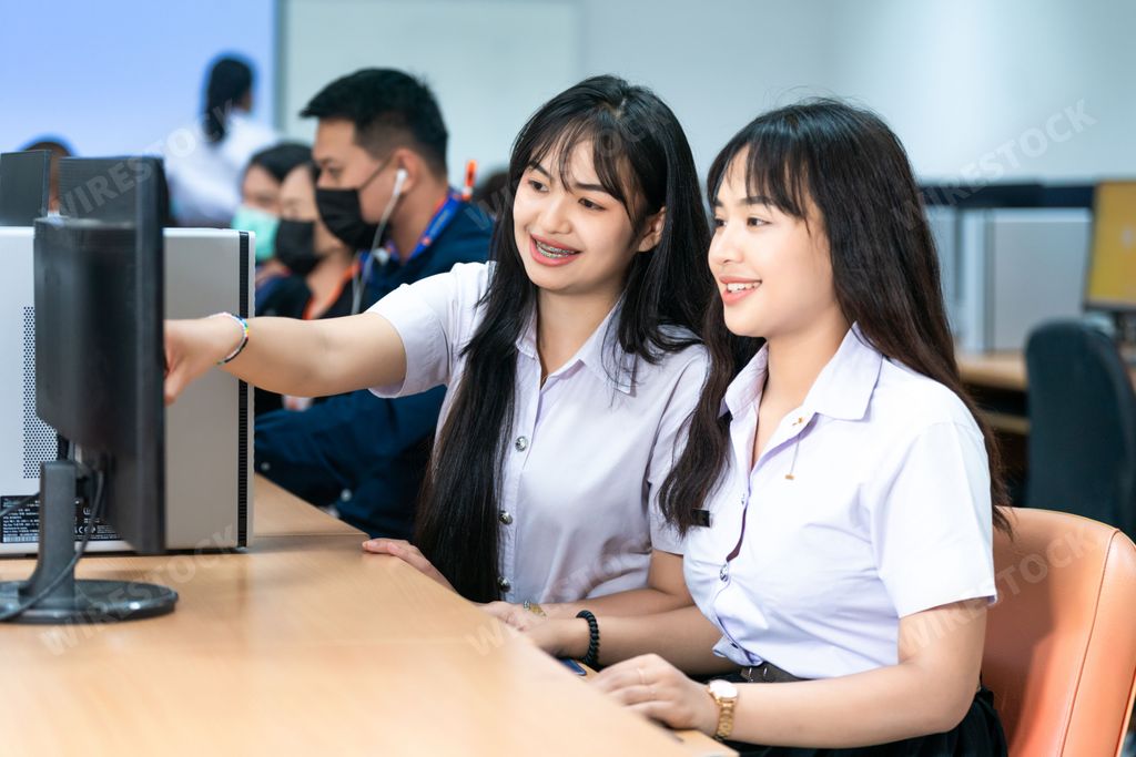 Two young Asian female students are seated at a desk, interacting with a laptop computer