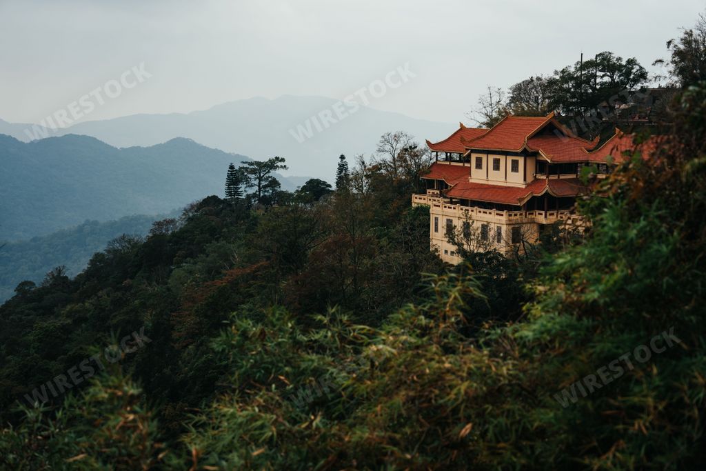 A two-story house with a red-tiled roof resting atop a mountain peak, surrounded by tall pine trees and lush vegetation