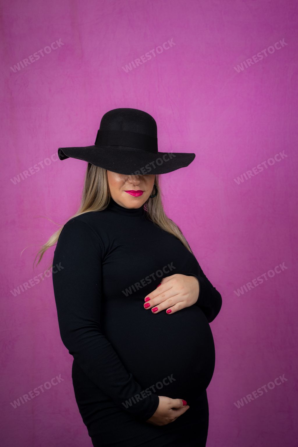A pregnant Caucasian woman posing in a black dress for a maternity shoot on a pink background