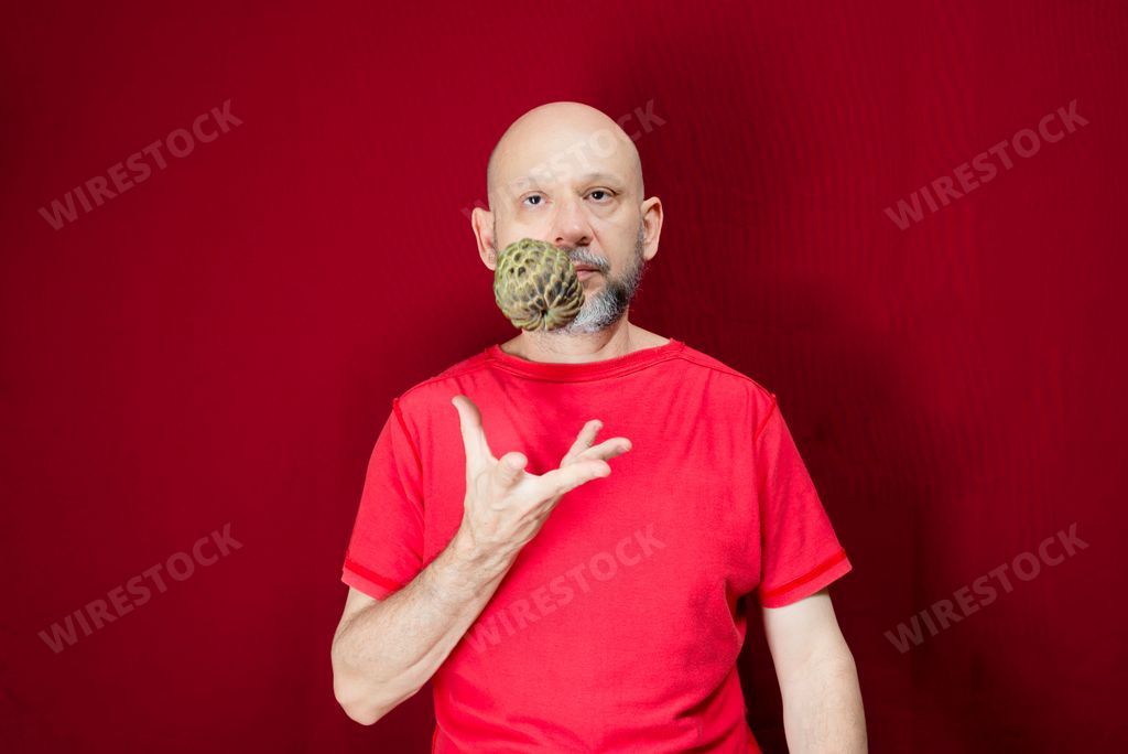 Young handsome man with beard, bald head and red shirt standing tossing pinecone fruit up against red background. Positive and healthy person.