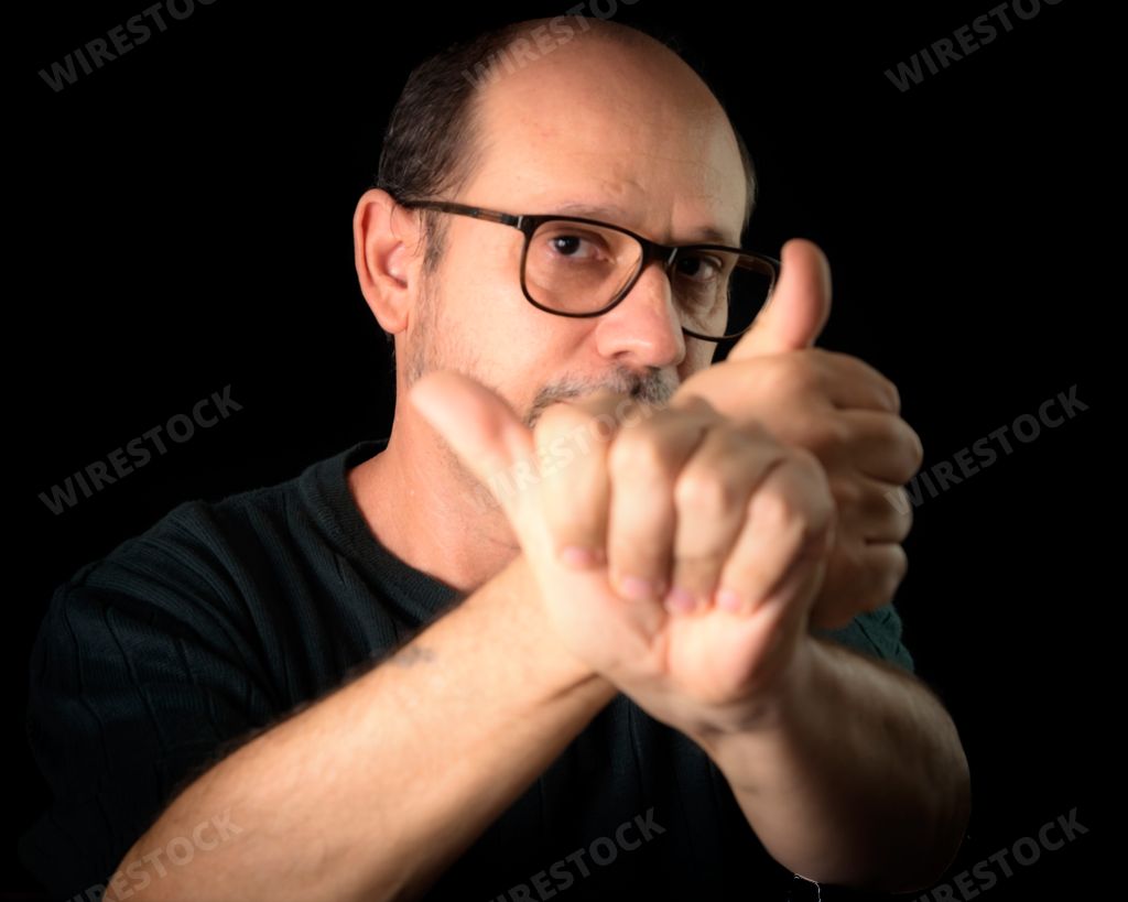 A closeup of a bearded man wearing glasses and gesturing with his hands against a black background