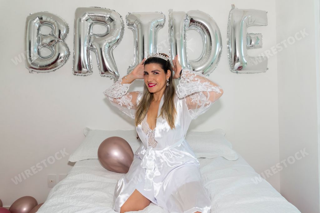 A young happy Brazilian woman in a white silk robe sitting on a bed in front of a Bride balloon at Bachelorette party
